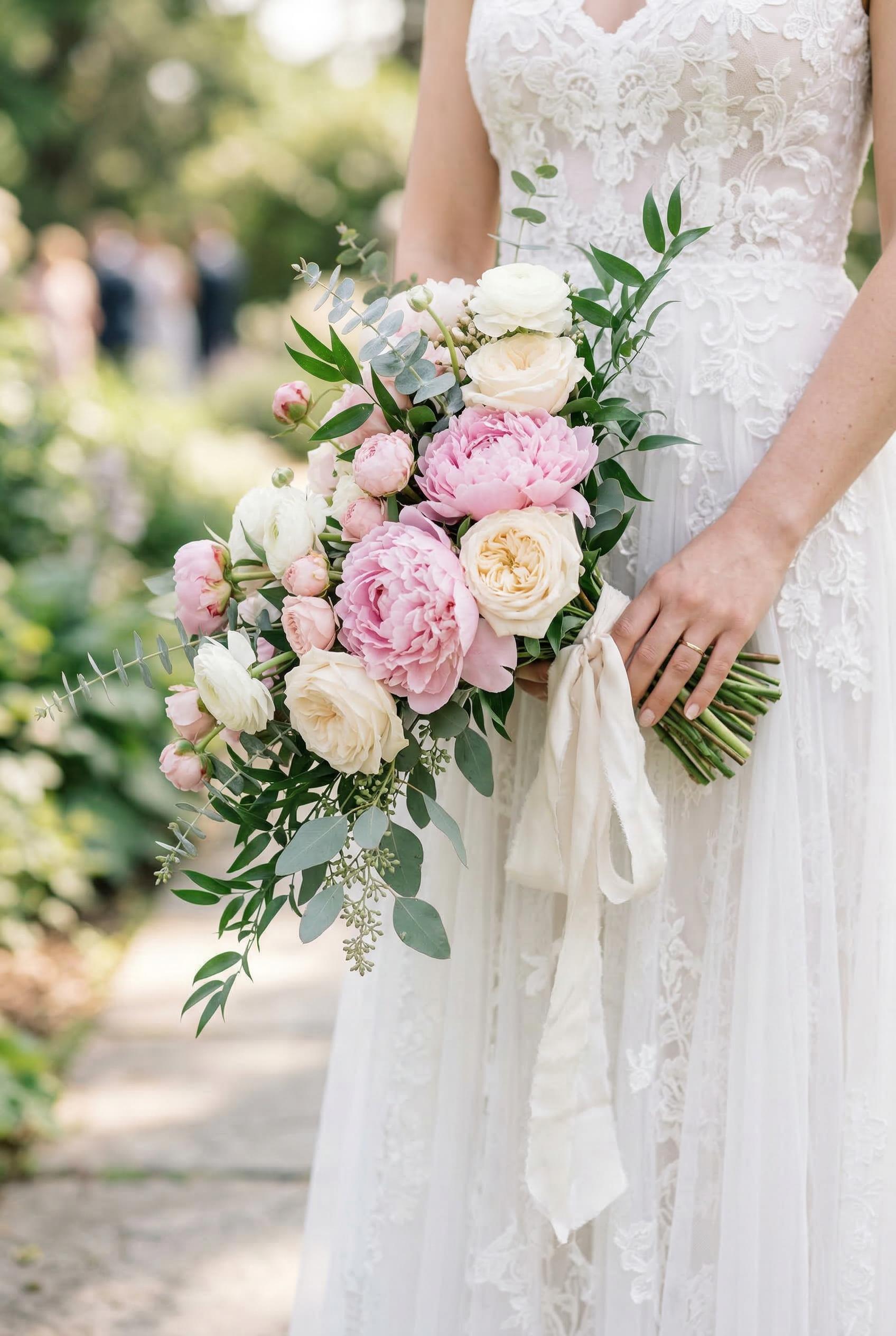 A romantic bridal bouquet of soft pink peonies, cream garden roses, white ranunculus, trailing eucalyptus and Italian ru