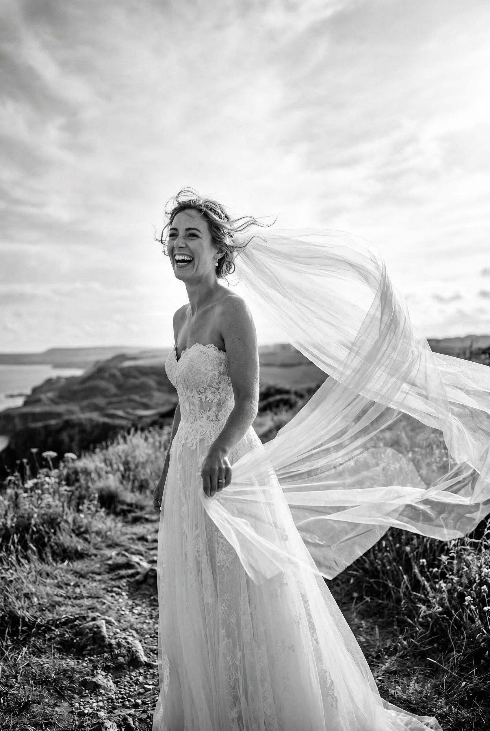 A striking black and white photograph of a bride laughing candidly while holding her long tulle veil
