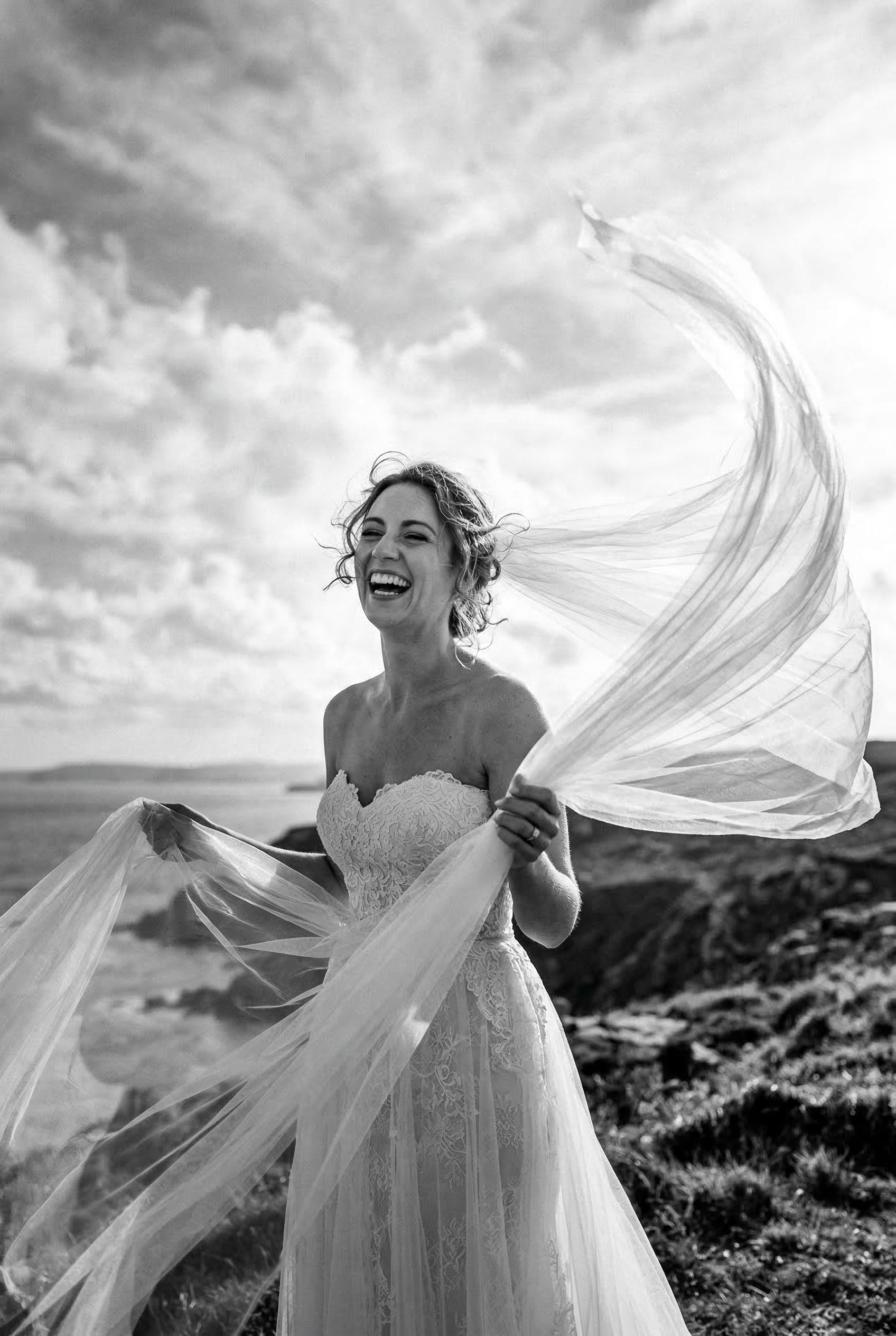 A striking black and white photograph of a bride laughing candidly while holding her long tulle veil as it billows drama