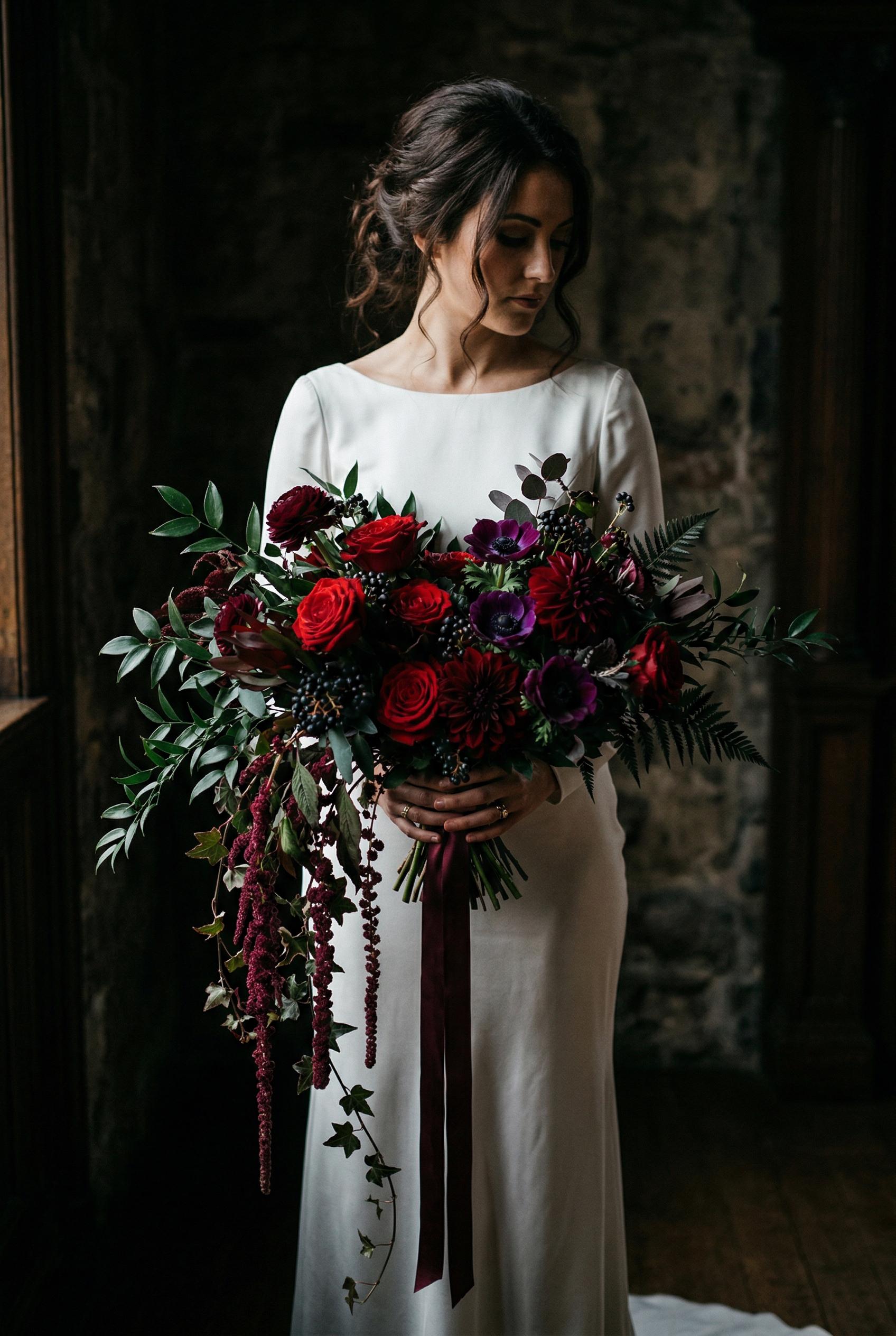 A dramatic oversized cascading bridal bouquet featuring deep crimson red roses, rich burgundy dahlias, dark purple anemo