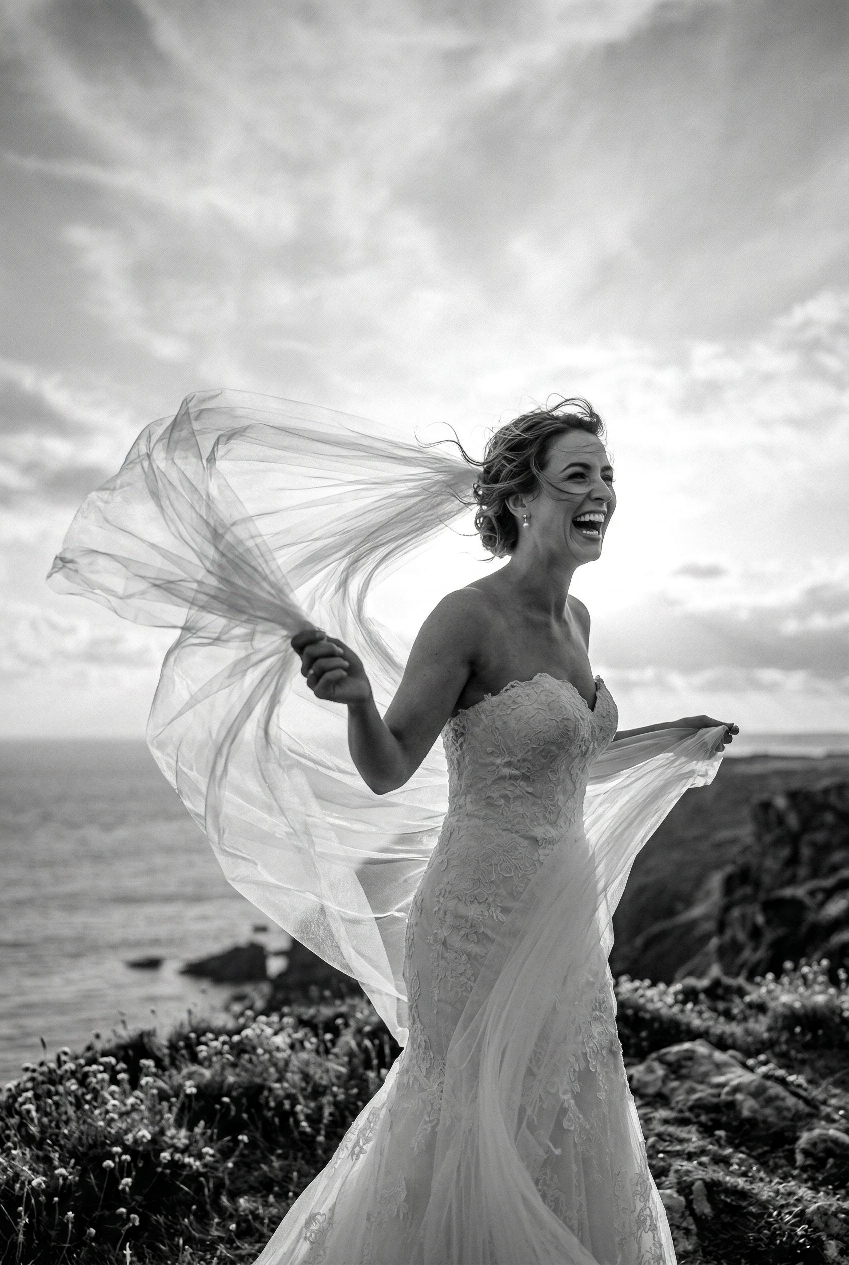 A striking black and white photograph of a bride laughing candidly while holding her long tulle veil as it billows drama