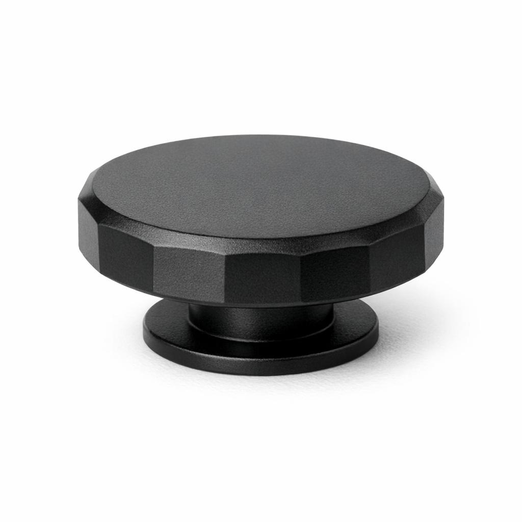 Commercial product photograph. A single residential cabinet knob. Faceted surface texture. Round Knob shape. Matte Black metal finish. Small circular base plate, single mounting post, approximately 1-1/4 inch diameter. Photographed on pure white seamless background (#FFFFFF). Studio softbox lighting, soft shadow directly underneath only. Macro lens, sharp focus throughout. Photorealistic real metal hardware. Not a 3D render. Not an illustration. Not CGI. Real cabinet hardware product photography quality. Variation 2: Paxton style cabinet knob: flat low puck shape with faceted angular sides cut into flat planes around the rim, flat top, thin base ring..