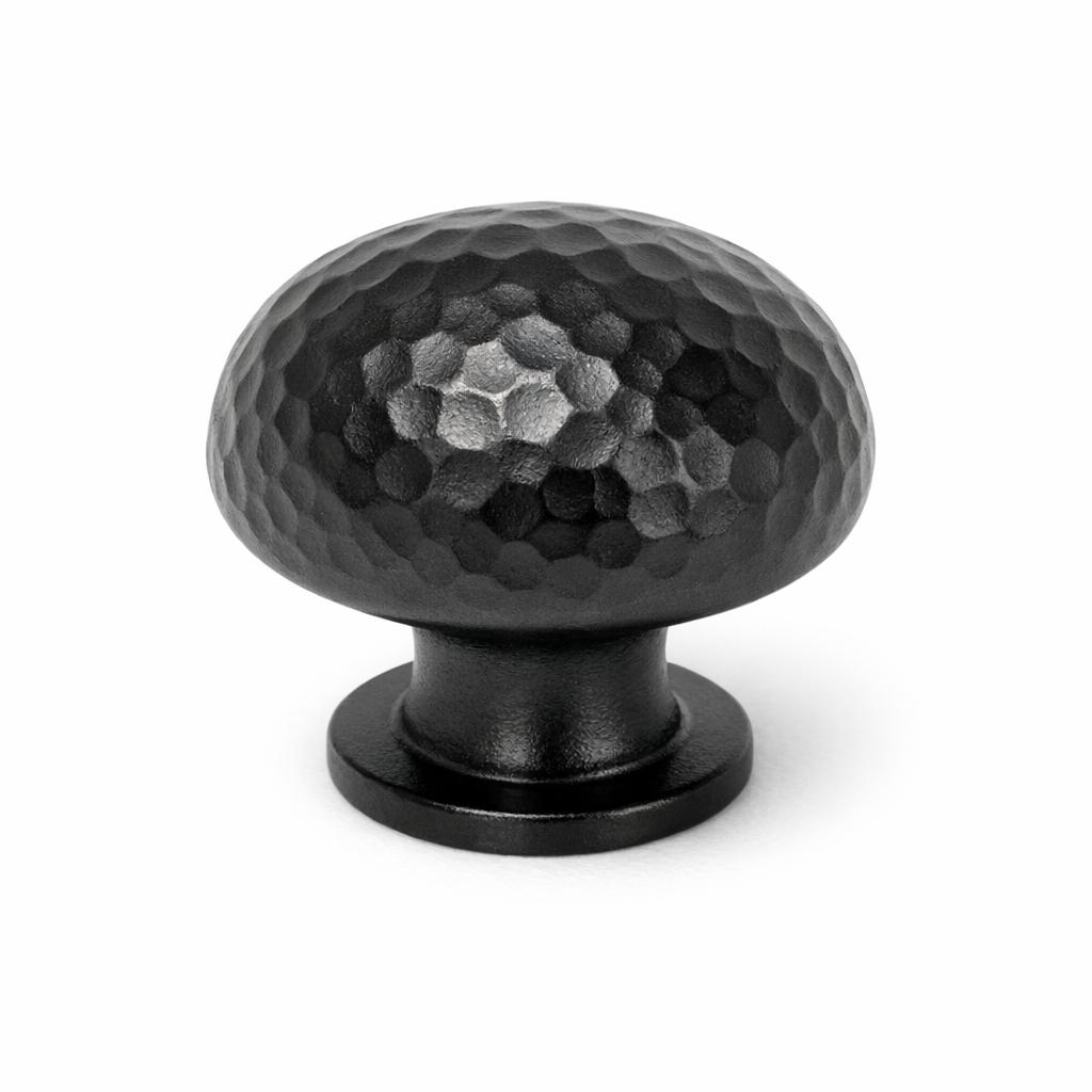 Commercial product photograph. A single residential cabinet knob. Hammered surface texture. Round Knob shape. Matte Black metal finish. Small circular base plate, single mounting post, approximately 1-1/4 inch diameter. Photographed on pure white seamless background (#FFFFFF). Studio softbox lighting, soft shadow directly underneath only. Macro lens, sharp focus throughout. Photorealistic real metal hardware. Not a 3D render. Not an illustration. Not CGI. Real cabinet hardware product photography quality. Variation 1: Providence style cabinet knob: tall rounded mushroom dome, hand-hammered planished texture across the entire dome with small rounded dimples catching the light, short thick neck, small circular base plate..
