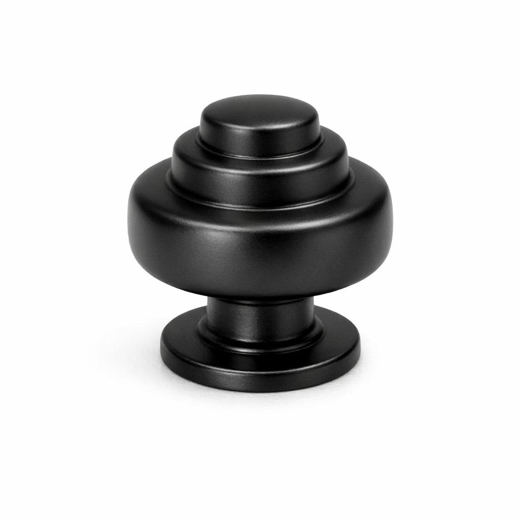 Commercial product photograph. A single residential cabinet knob. Smooth surface texture. Round Knob shape. Matte Black metal finish. Small circular base plate, single mounting post, approximately 1-1/4 inch diameter. Photographed on pure white seamless background (#FFFFFF). Studio softbox lighting, soft shadow directly underneath only. Macro lens, sharp focus throughout. Photorealistic real metal hardware. Not a 3D render. Not an illustration. Not CGI. Real cabinet hardware product photography quality. Variation 7: Tiered knob: stacked cylindrical tiers getting smaller toward top, like a small wedding cake shape, circular base. Art deco style..